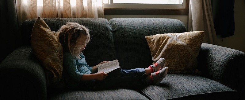 Niña rubia sentada en un sofá lee un libro con poca luz 