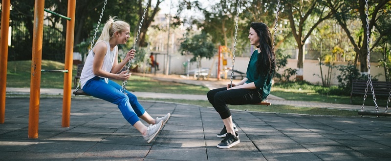 Dos mujeres jóvenes se ríen sentadas en los columpios de un parque