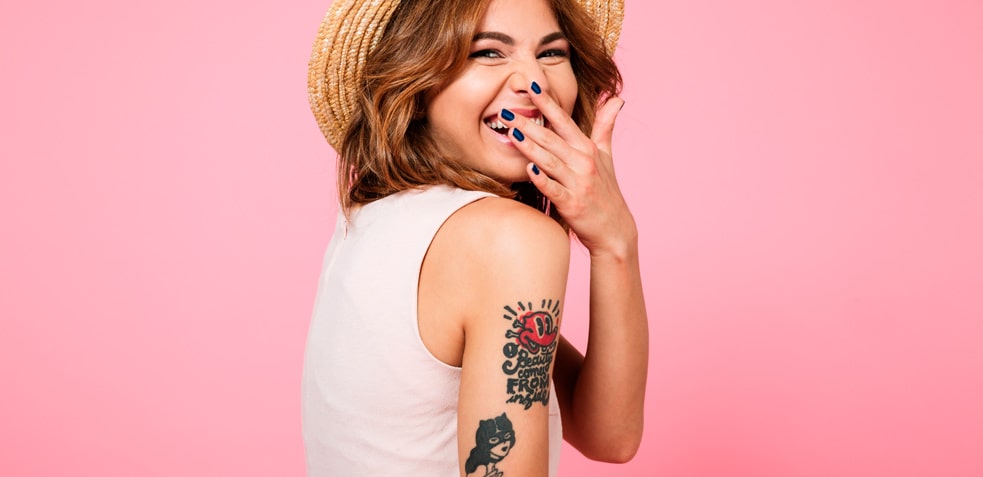 Mujer con sombrero sonríe y se tapa la boca con su mano mostrando los tatuajes de su brazo 