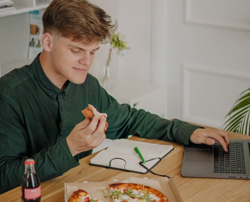 Adolescente con camisa verde come una pizza mientras utiliza un ordenador portátil
